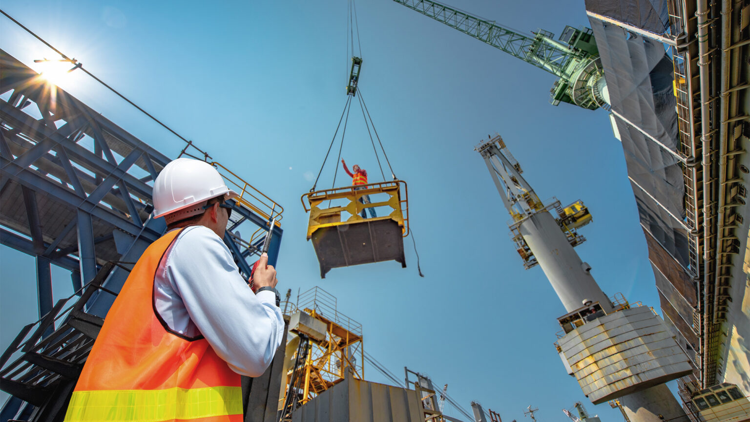 Worker looking at a cellphone with construction software while crane lifts another worker on a platform